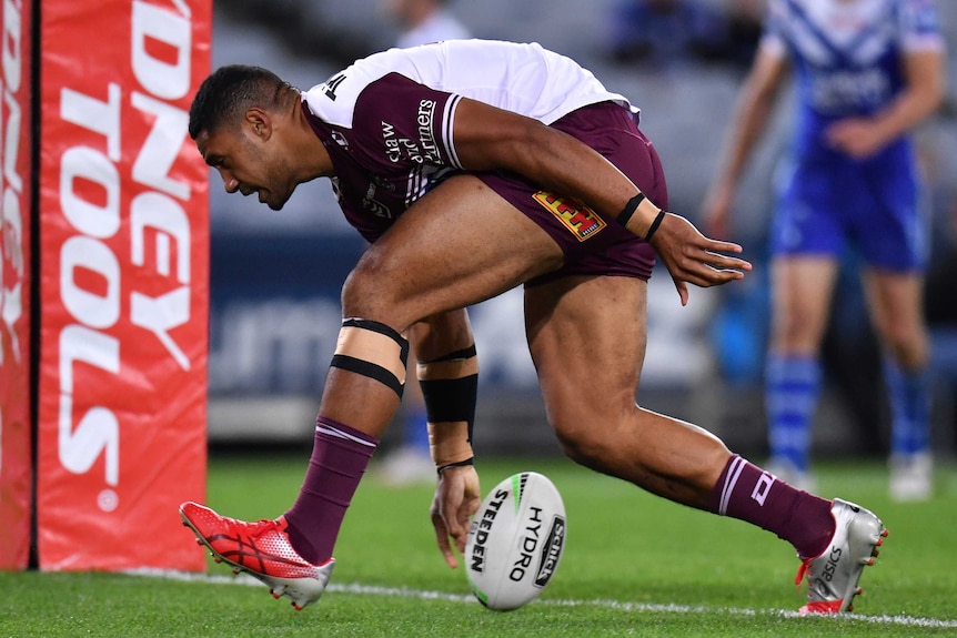 A Manly Sea Eagles NRL player puts the ball down on the ground with his right hand as he scores a try next to the goal posts.