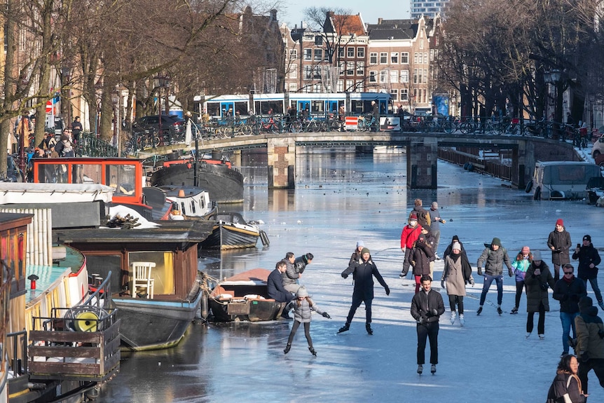 People ice skate on a frozen canal in front of boats and a bridge in the background
