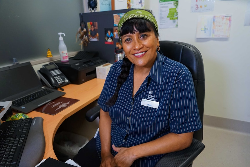 Heni Hongara sits at her desk and smiles at the camera. She is wearing a navy nurses uniform and a green bandana.