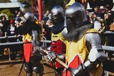 Three people in steel armour, holding Medieval weapons, prepare to battle as a crowd watches on