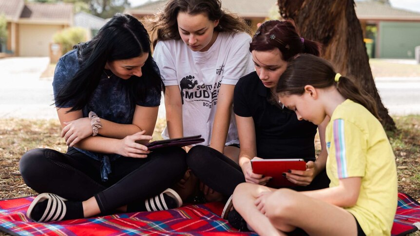 Four teenage girls sit on a picnic blanket in front garden with ipads