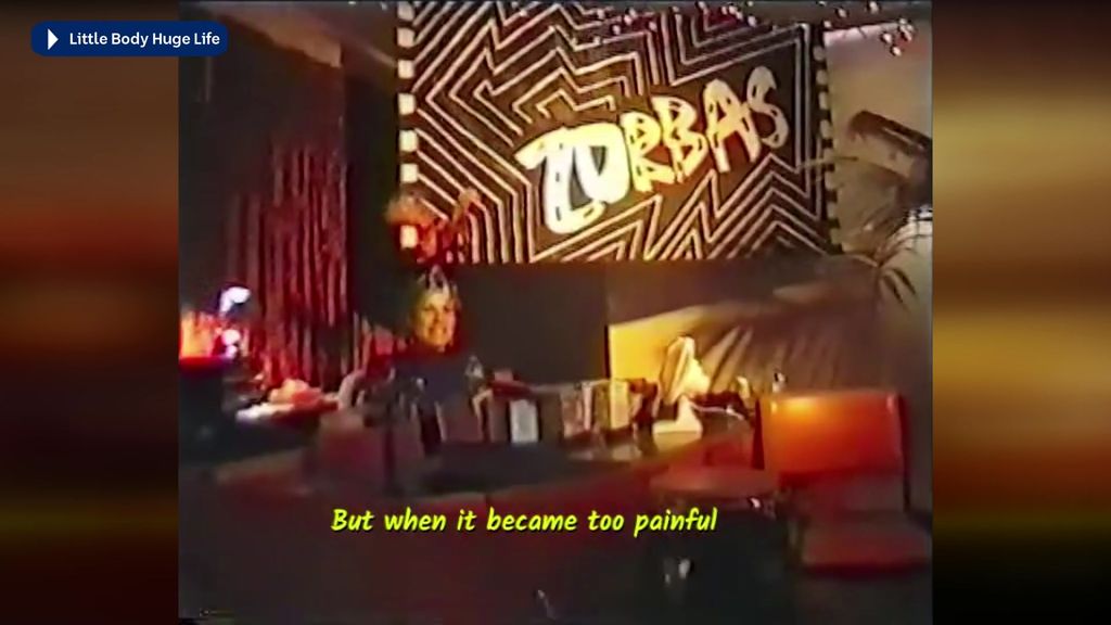 Archival footage of a woman behind a bar with a sign saying 'Zorbas' behind her. Subtitles say 'But when it became too painful'.