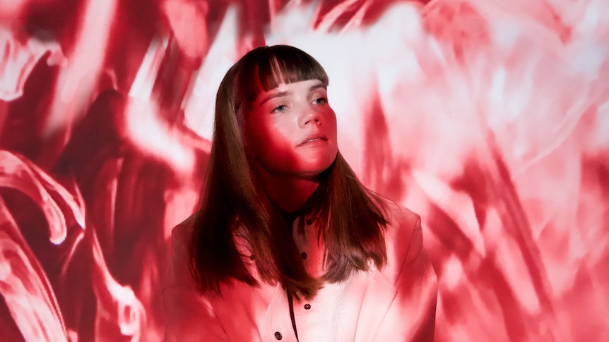 A woman with long hair and bangs looks thoughtfully to the side, while patterned pink and white lighting over and behind her