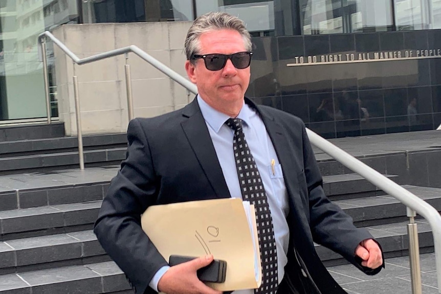 A man in a suit walks down steps outside court carrying a manila folder of papers.