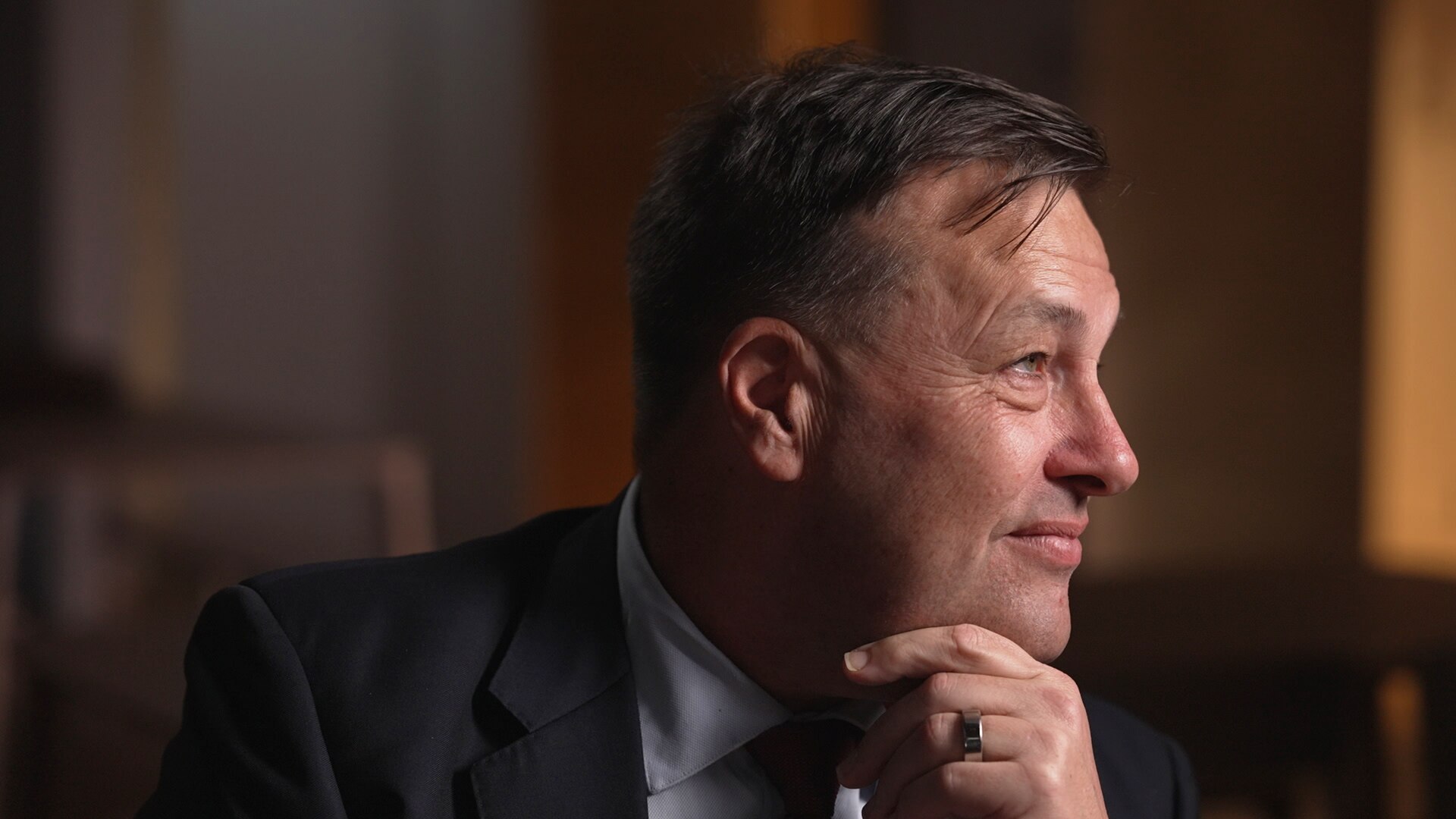 A man rests his head on his chin as he looks to his side in a dimly-lit room.