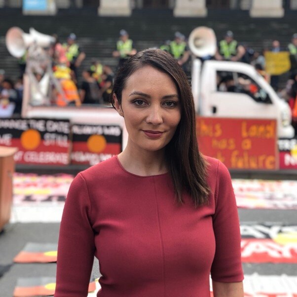 Head shot of a woman with long, brown hair standing in front of a protest with Indigenous flags on a truck.