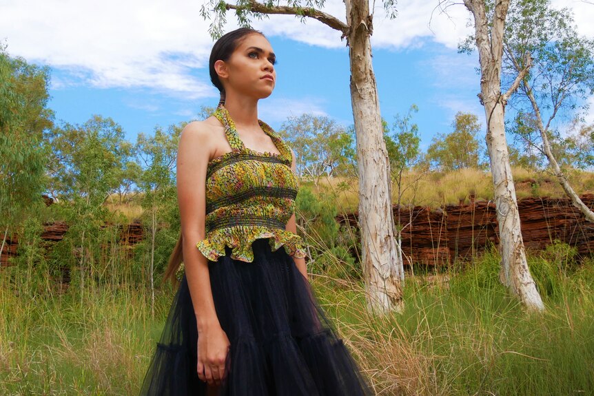 A young woman models a strappy green top and long black skirt amid tall grass, trees and red rocks. 