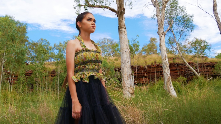 A young woman models a strappy green top and long black skirt amid tall grass, trees and red rocks. 