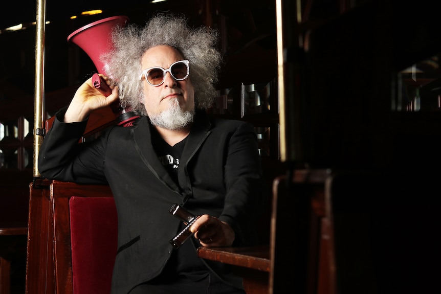 Sxip Shirey sits at a Spiegeltent booth holding a megaphone and harmonicas.