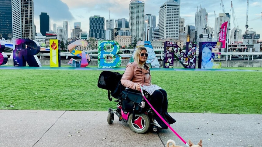 A white woman in an electric wheelchair is walking her dog with a pink leash. She's in front of green grass and a BRISBANE sign