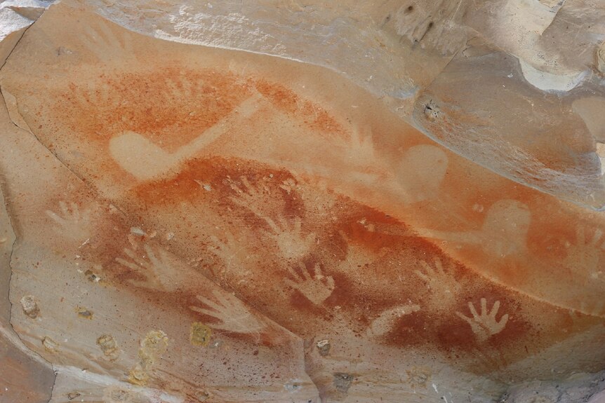 Ancient Indigenous hand art in rock with many hand prints shown up against an orange colour in the rock.