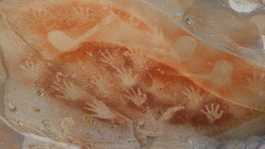 Ancient Indigenous hand art in rock with many hand prints shown up against an orange colour in the rock.