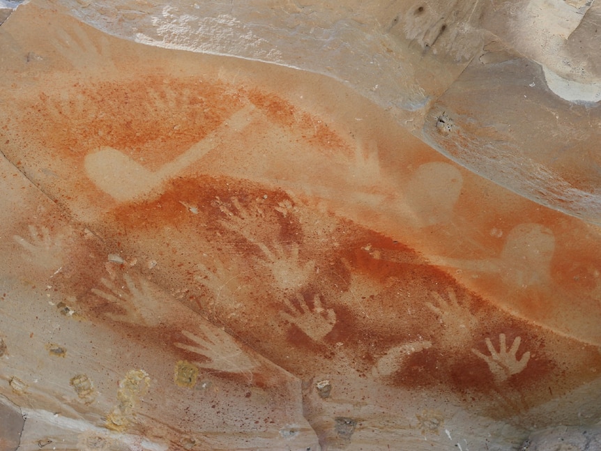 Ancient Indigenous hand art in rock with many hand prints shown up against an orange colour in the rock.