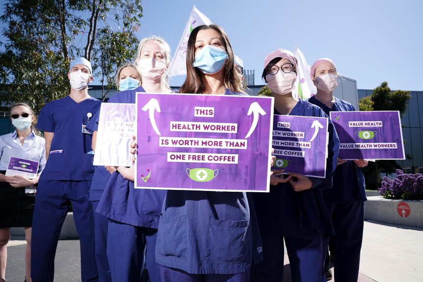 A group of people wearing blue medical scrubs and face masks hold protest signs