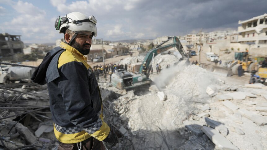 A volunteer in a white helmets stands at the site of damaged buildings as heavy equipment works to clear rubble.