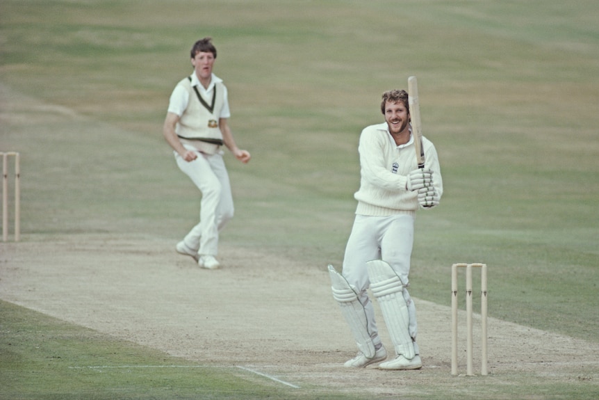 Ian Botham smiles while  holding a cricket bat with a bowler in the background