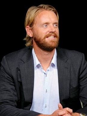 A man with orange hair and a beard smiles looking to the side