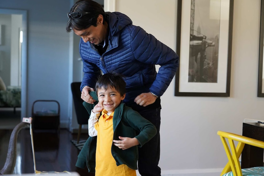 A young boy smiling and putting his school jumper on stands alongside his dad as they get ready for school.