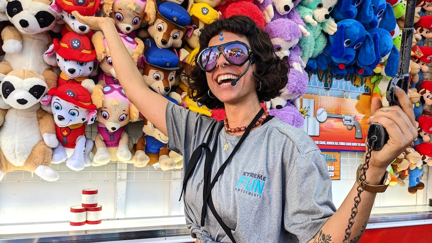 A trendy woman wearing sunglasses and a microphone headset gestures at a brightly coloured assortment of plush toy animals.