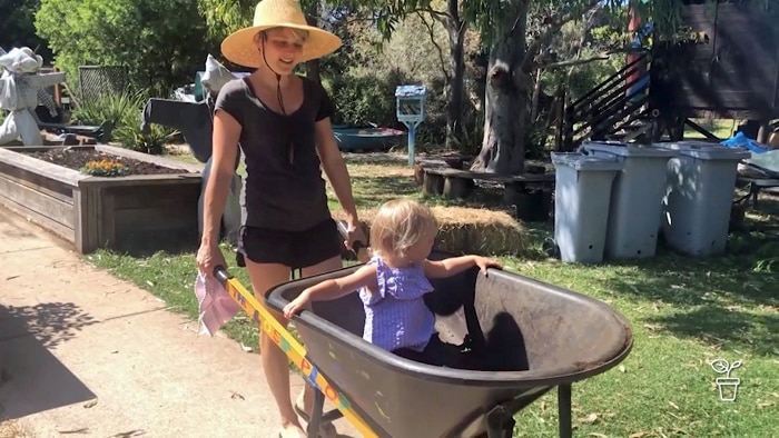 Woman in hat wheeling small child in wheelbarrow through garden