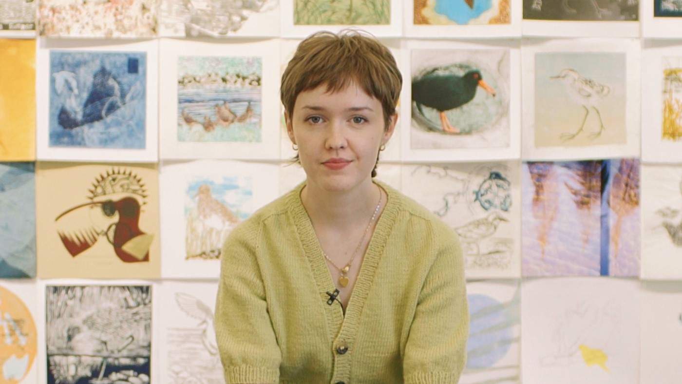 A woman in her mid 20s with short brown hair and wearing a light cardigan sits in front of a wall of drawings