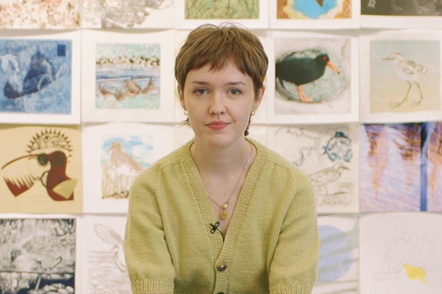 A woman in her mid 20s with short brown hair and wearing a light cardigan sits in front of a wall of drawings