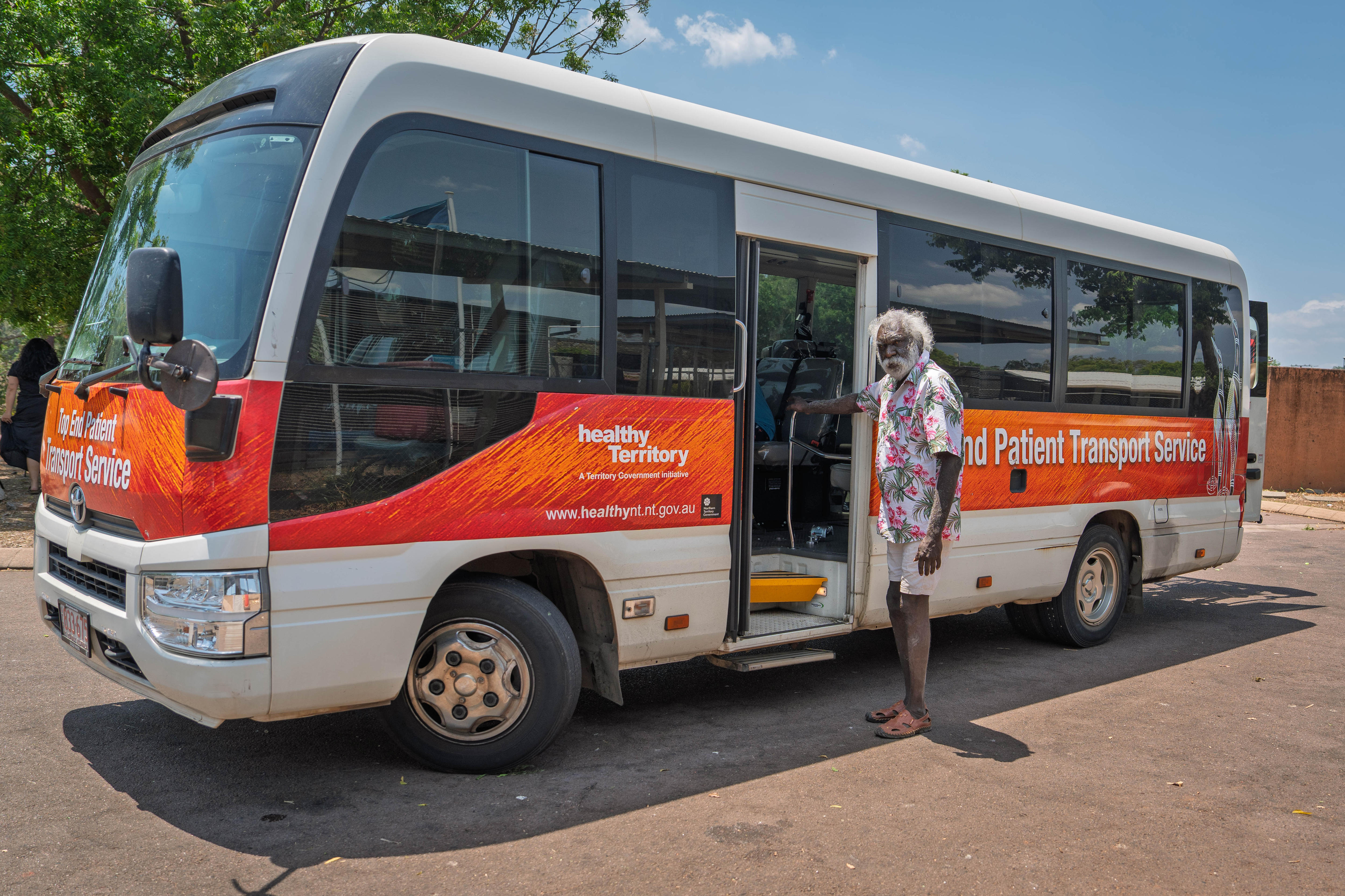 Charlie, an older Indigenous man, standing alongside a shuttle bus used to transfer medical patients in the Top End.