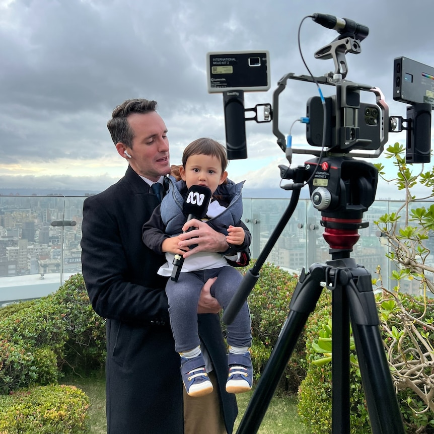 Man holding a toddler holding an ABC microphone standing in front of a camera.