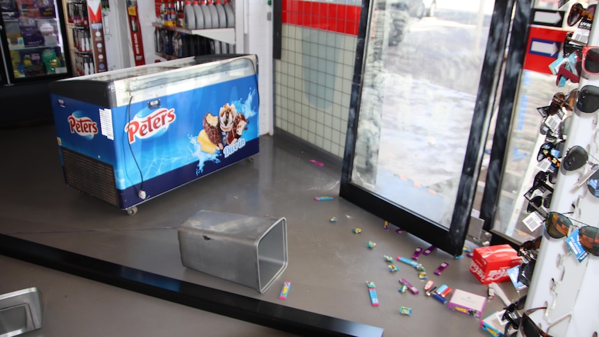 the floor of a business with a broken door and a bin, lollies and door parts scattered over the ground 