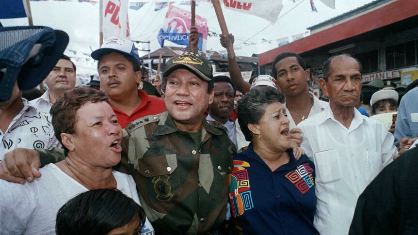 Manuel Noriega in army fatigues walks alongside cheering supporters waving signs in Panama City. The photo is from 1989.