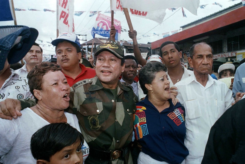 Manuel Noriega in army fatigues walks alongside cheering supporters waving signs in Panama City. The photo is from 1989.