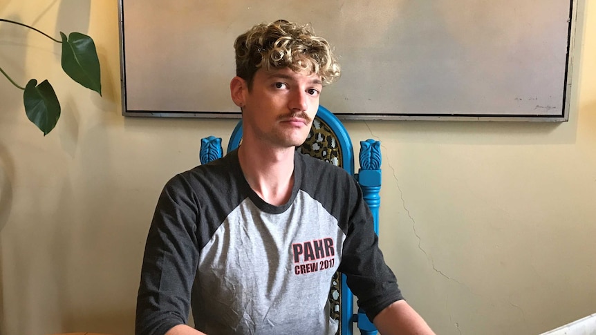 A young man sitting at a table in front of a laptop looking sadly at the camera