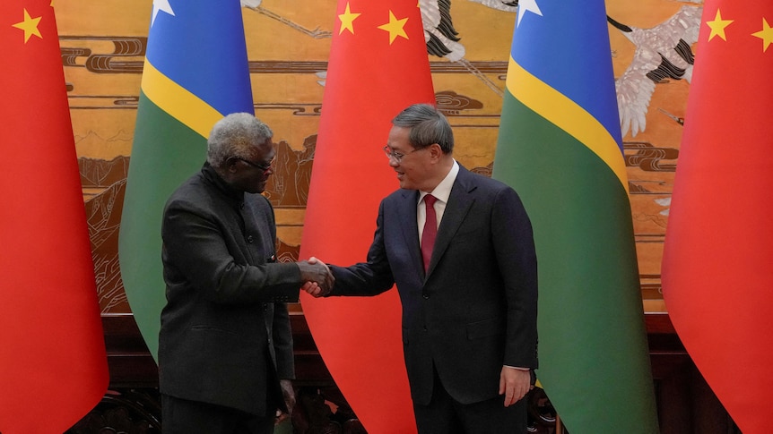 Manasseh Sogavare shakes hands with his Chinese counterpart Li Qiang in front of flags from both of their countries.