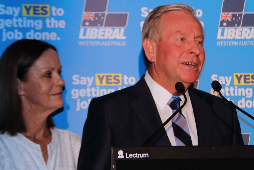 A mid shot of Colin Barnett talking on stage in front of his wife and a Liberal backdrop.