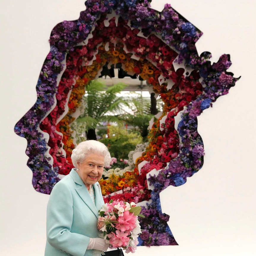 Queen Elizabeth II stands in front of a floral exhibit featuring a profile image of herself.
