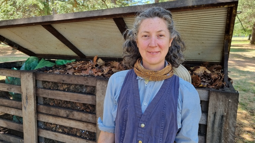 A woman with grey hair stands in front of a large compost heap.