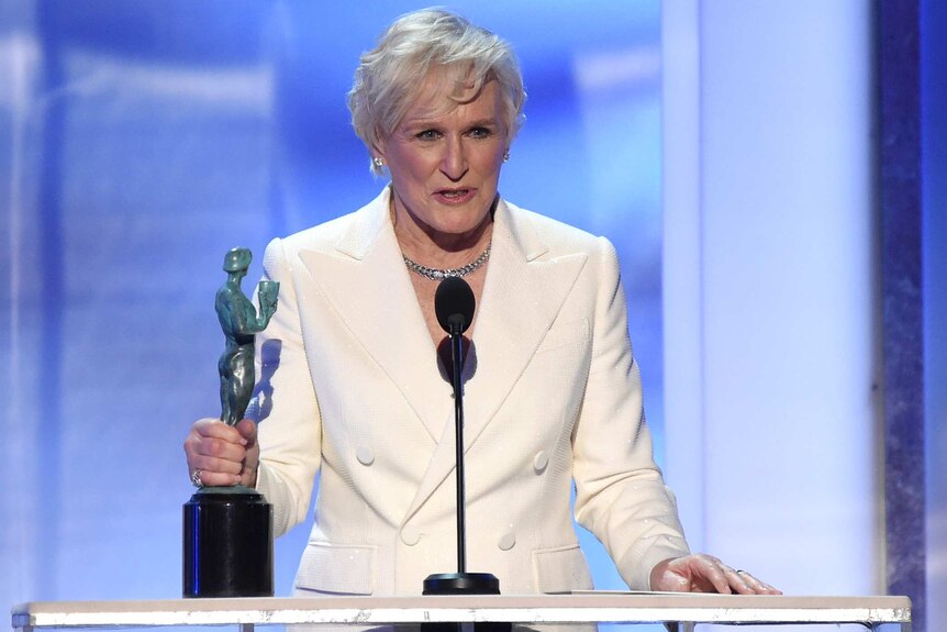 Glenn Close wears a white suit as she stands at podium with SAG award in her hand while delivering speech.