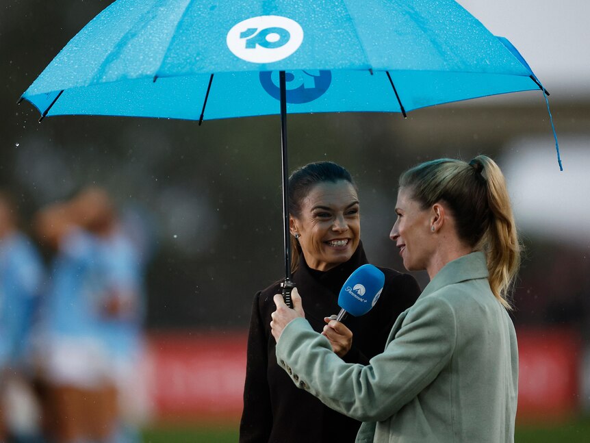 Two women wearing winter coats stand beneath a blue umbrella while speaking on live television