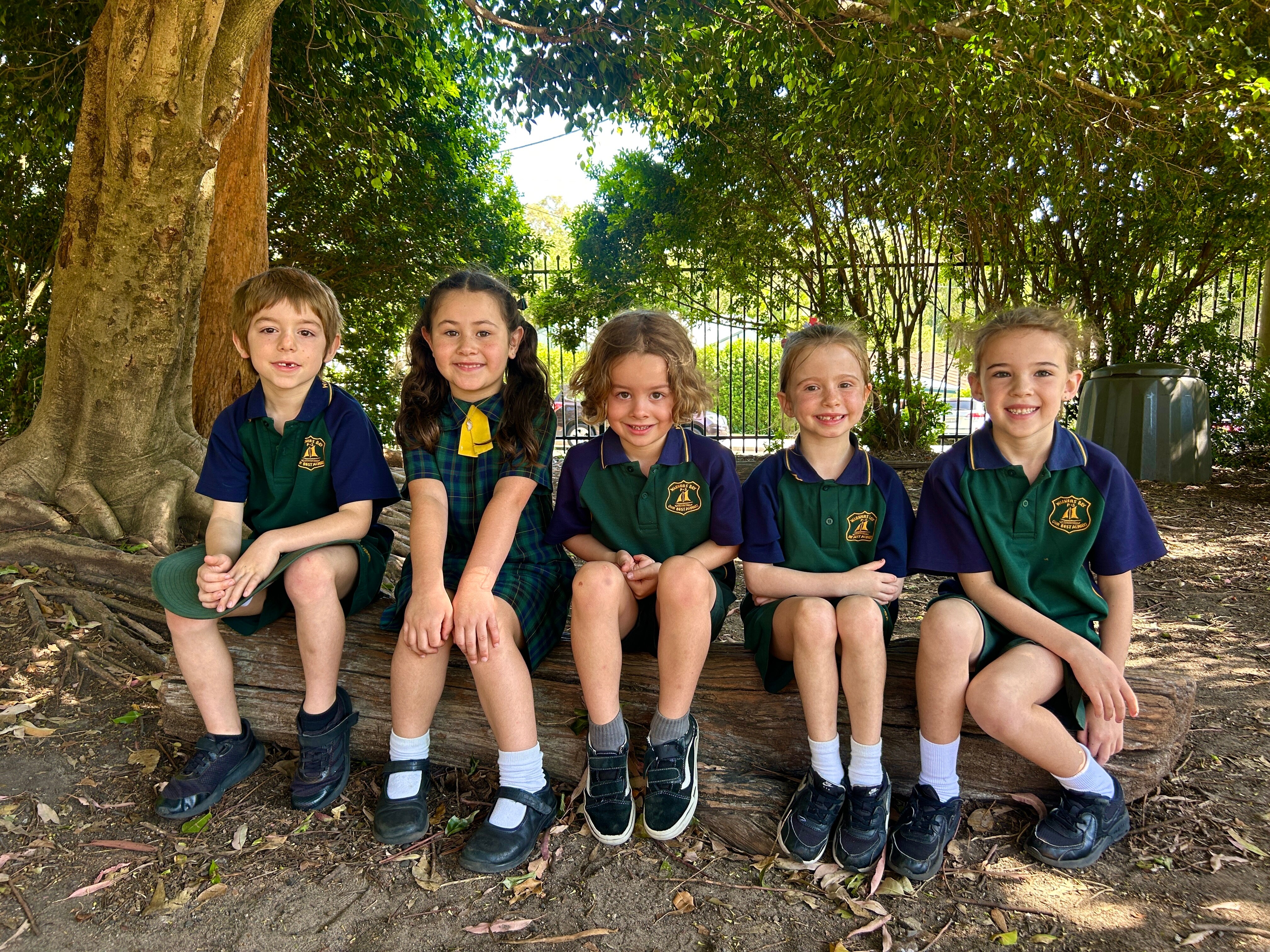 Five young children sit on a log smiling. They are wearing green school uniforms