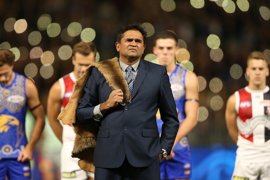 Nicky Winmar holds a pelt over his shoulder while standing in front of AFL players at a game between St Kilda and West Coast.
