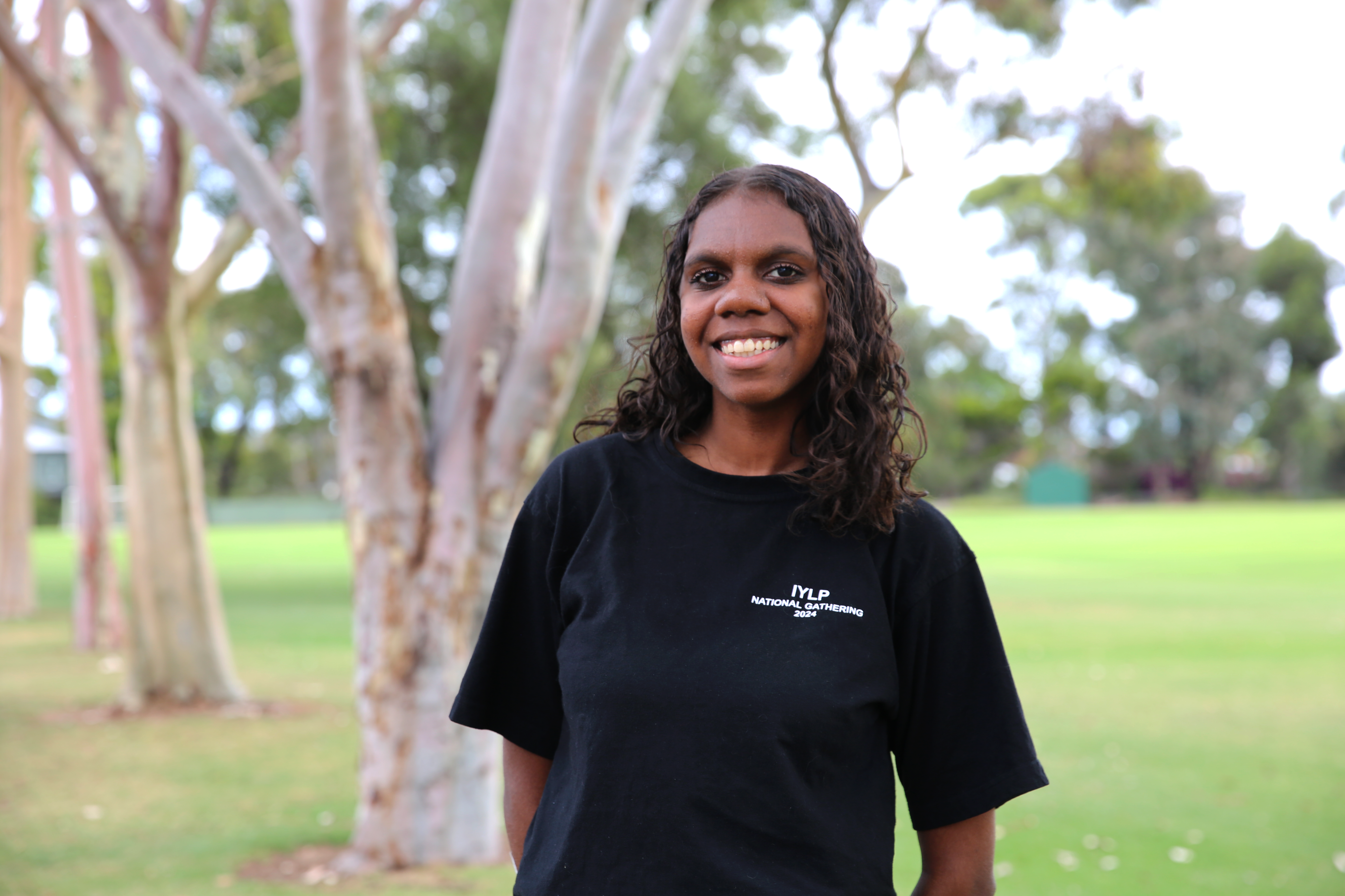 A young woman with black skin and brown hair smiles at the camera, she is outside in front of trees and grass
