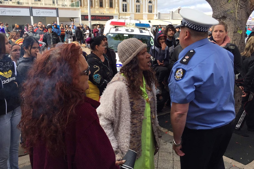 A woman confronts a police officer in a crowd of people with a police vehicle in the background.