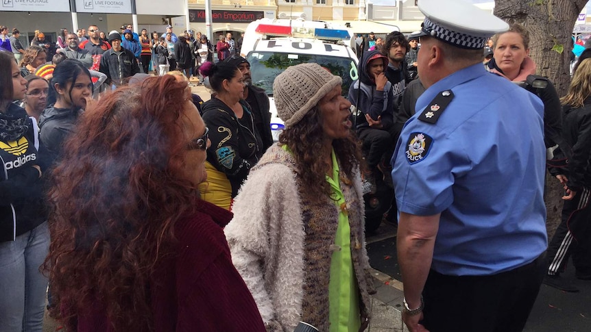 A woman confronts a police officer in a crowd of people with a police vehicle in the background.