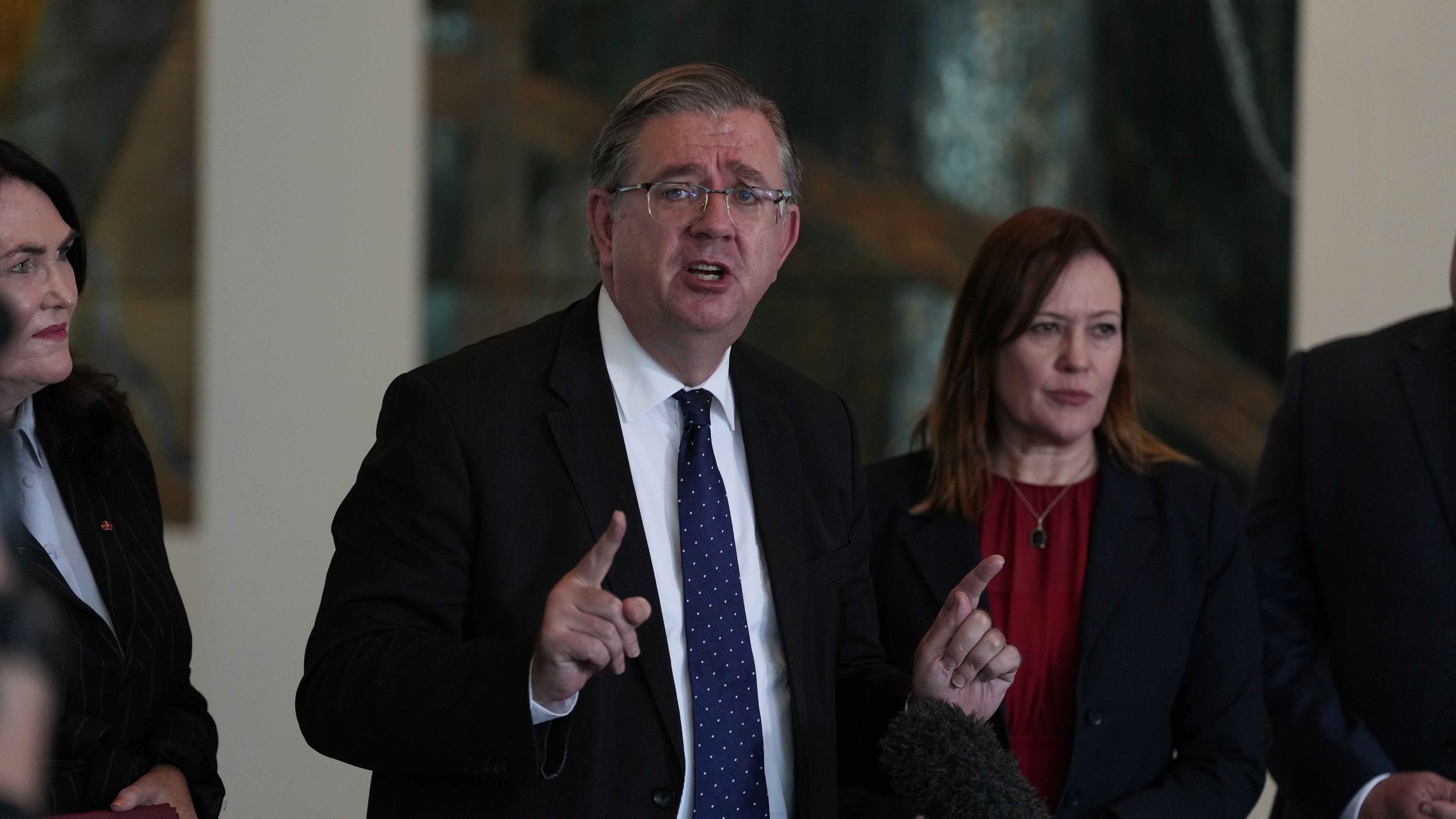 A middle-aged man in a dark suit gestures as he speaks.