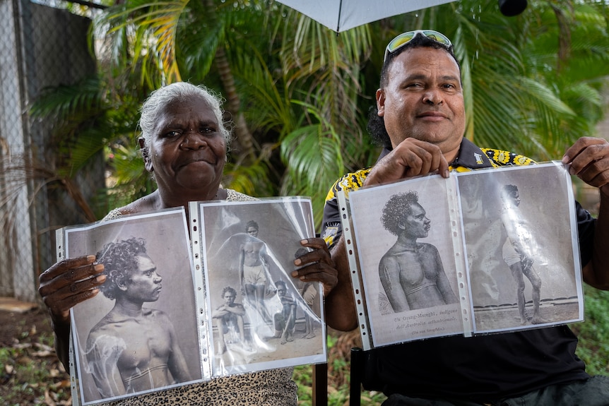 A woman and a man sit outside holding up old black and white photographs of young Aboriginal men
