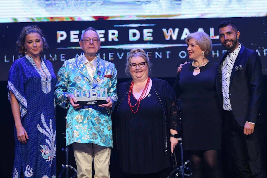 Men and women on a stage with one man holding a trophy shaped like the letters LGBTI