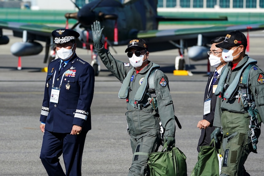 Moon Jae-in in military flying gear and a surgical mask waves from an asphalt