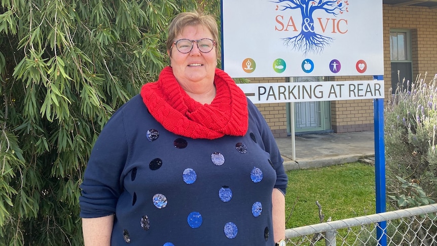 A woman with short hair, glasses, a red scarf and a blue polka-dot jumper stands smiling in front of a sign.