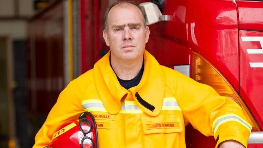 A firefighter wearing a yellow safety jacket and holding a hardhat leans on a firetruck with a serious expression.
