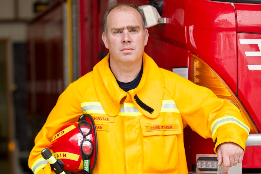 A firefighter wearing a yellow safety jacket and holding a hardhat leans on a firetruck with a serious expression.